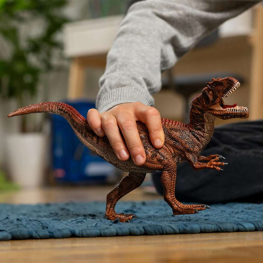 A child's hand holds a light brown with dark brown speckles Allosaurus plastic figurine on a blue carpet. The dinosaur's head is reared back, mouth open baring teeth, with short arms in front.